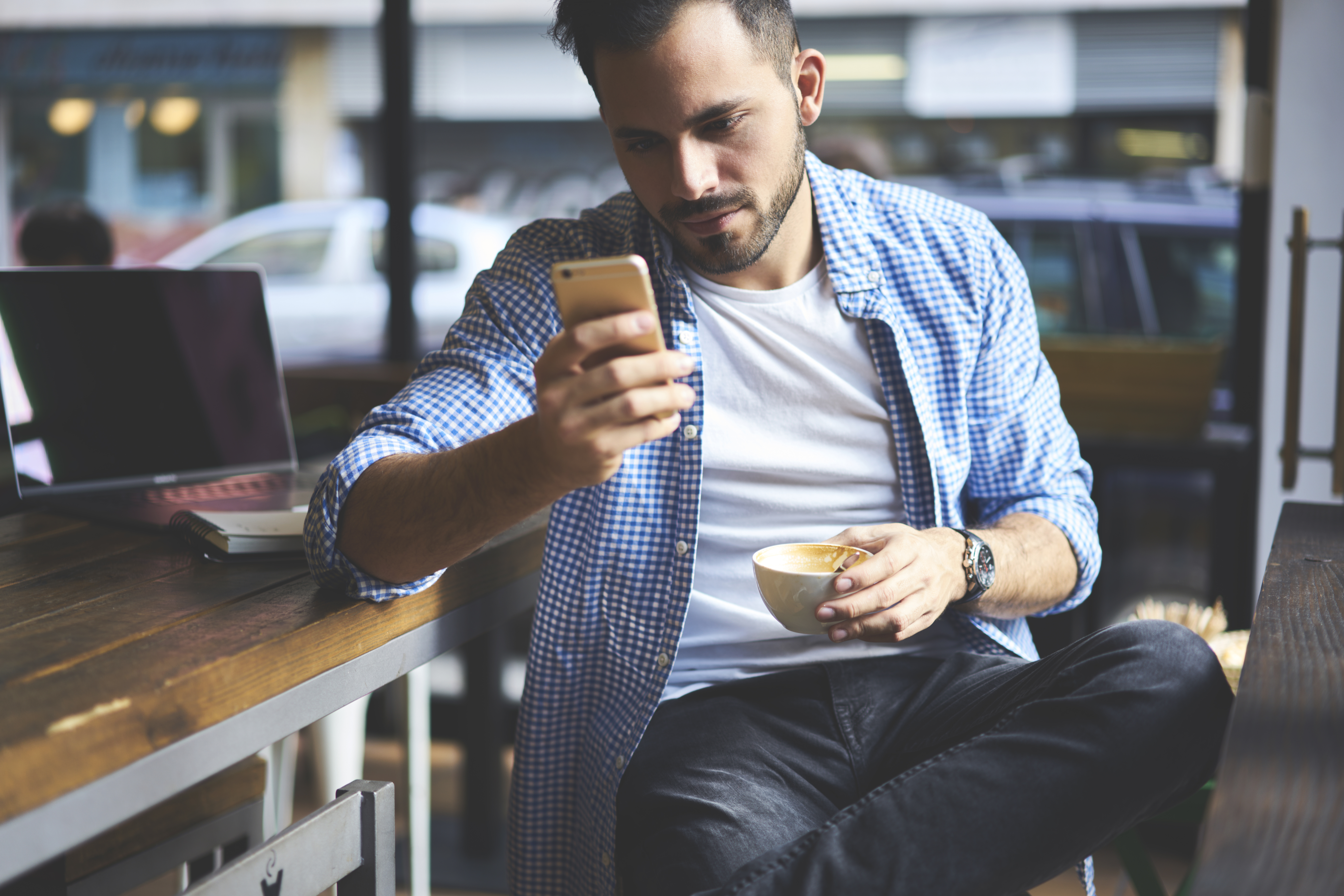 Concentrated casually dressed male freelancer checking email box waiting for transaction using online banking service, serious hipster guy installing application on smartphone during coffee break